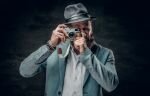 A man dressed in a grey jacket and felt hat holds an SLR photo c